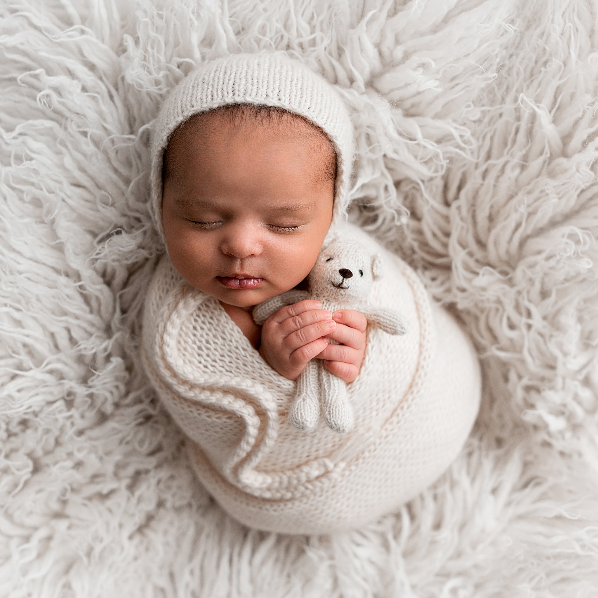 Newborn Photoshoot Wakefield. Sleeping newborn baby swaddled neatly in a cream knitted blanket and wearing a matching bonnet. He's nestled snug into a fluffy fur rug while holding a tiny teddy in his hands.