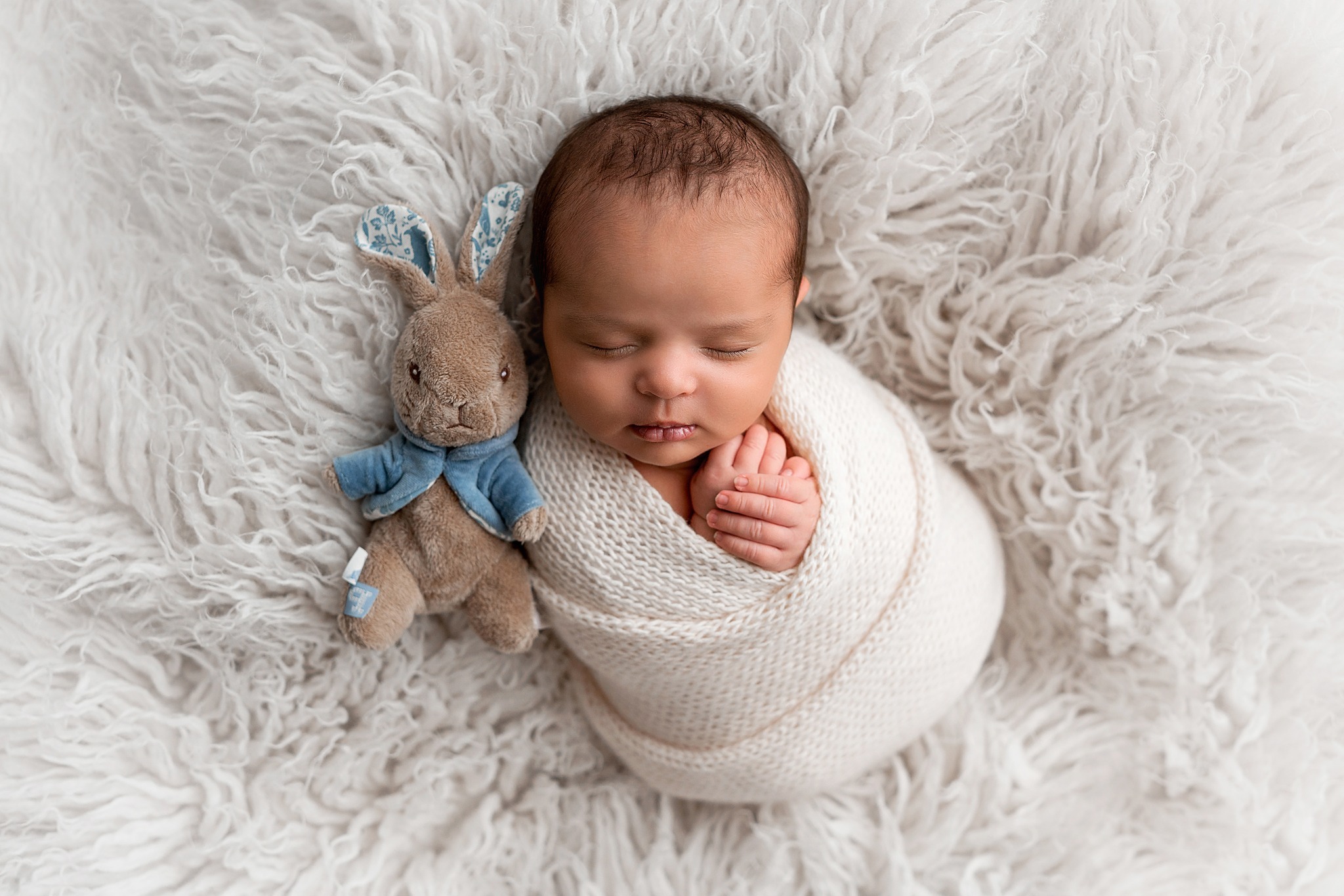 Newborn Photoshoot Wakefield. Sleeping newborn baby swaddled neatly in a cream knitted blanket. He's nestled snug into a fluffy fur rug and snuggled up to a mini Peter Rabbit cuddly toy.