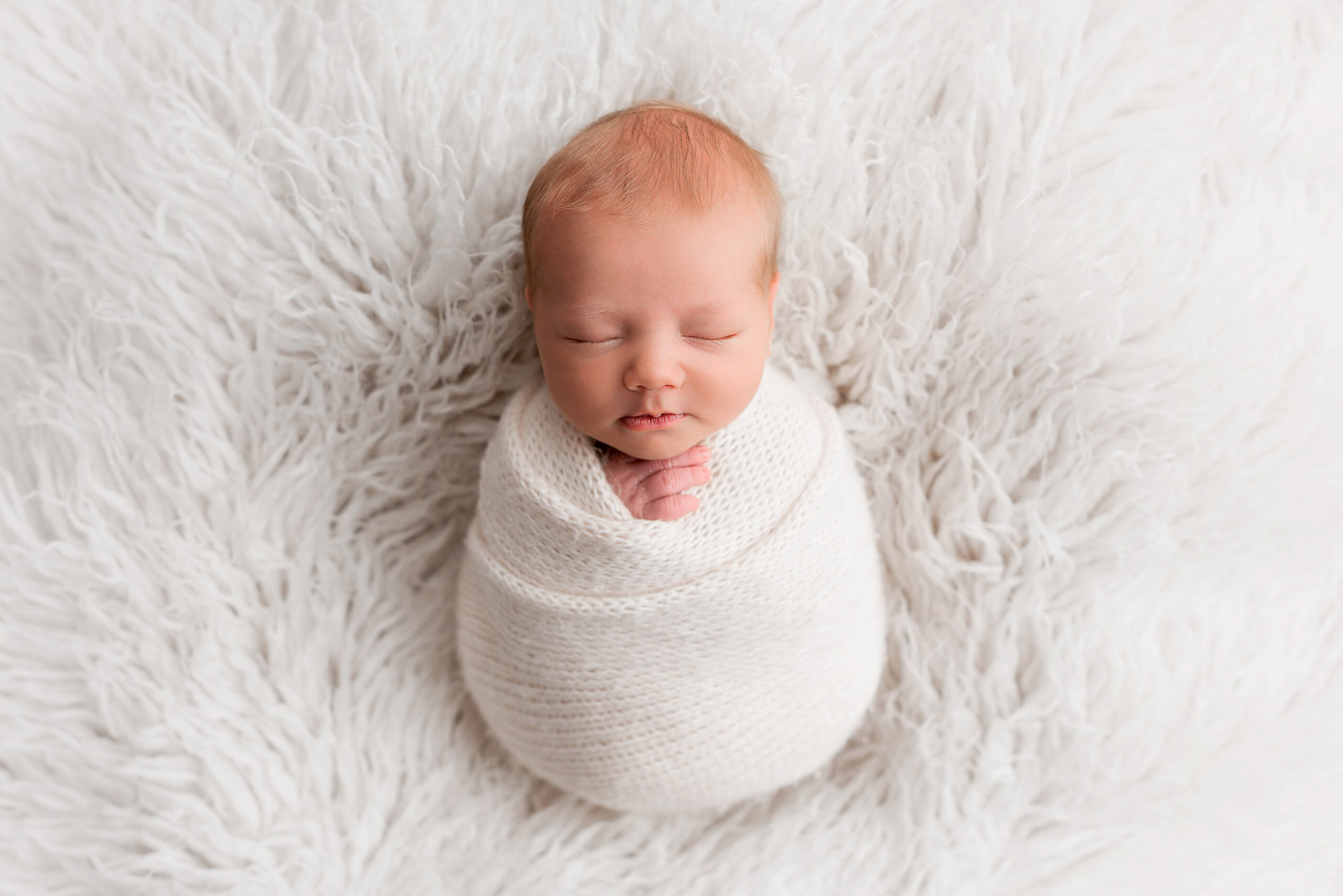 Newborn Photography Barnsley, newborn baby swaddled in a cream knitted blanket laid sleeping on a fluffy cream background.